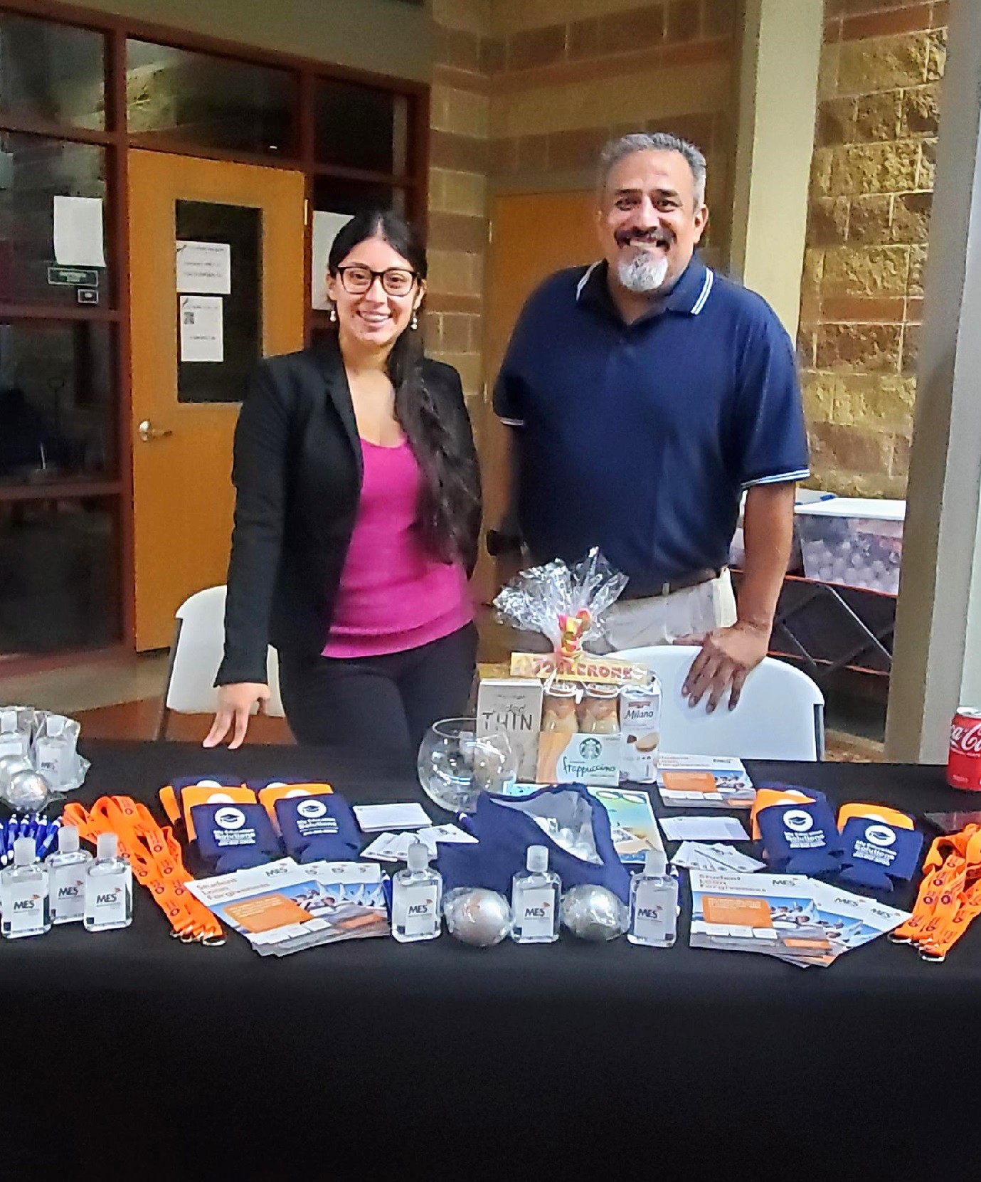 A woman and man stand behind a table filled with branded giveaway items greeting event attendees with a smile