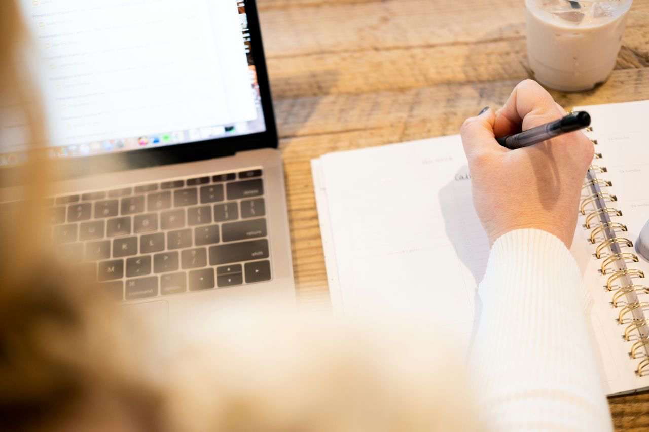 An overhead shot of a woman writing something on a pad of paper while looking at a laptop screen